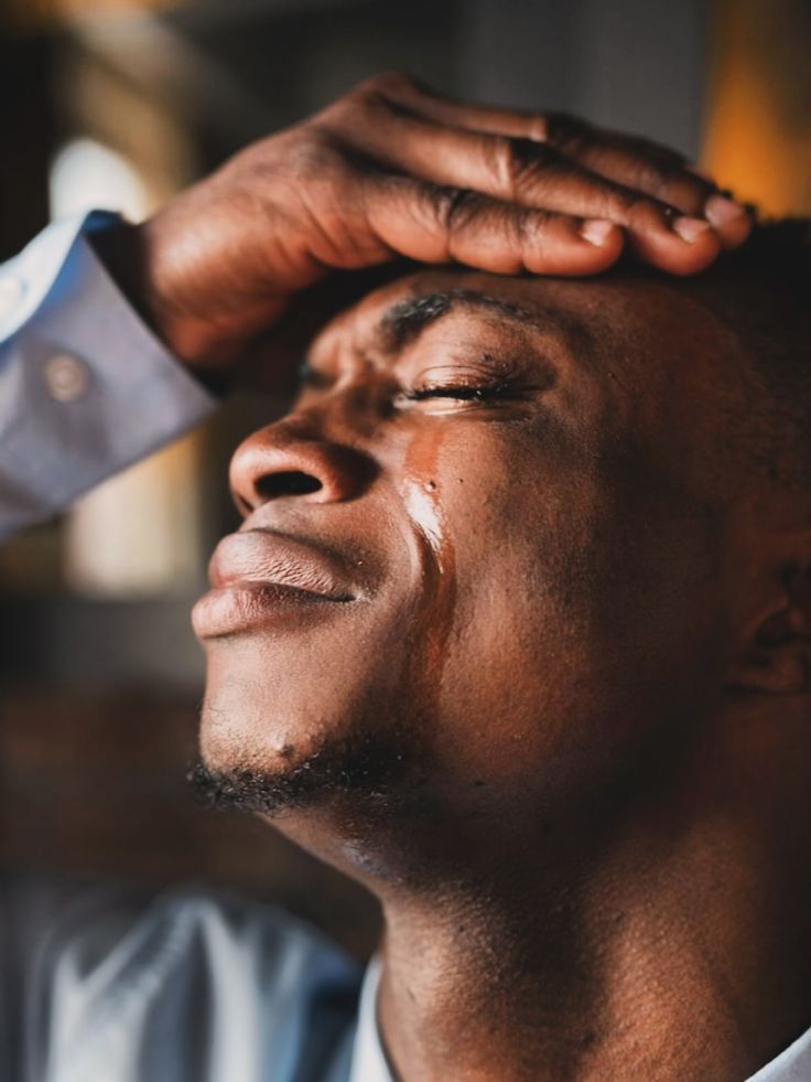 A Ghanaian man with eyes closed and a tear on his cheek, hand resting on his forehead