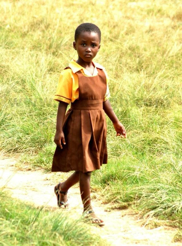 A Ghanaian schoolgirl in yellow and brown uniform walking a grass path
